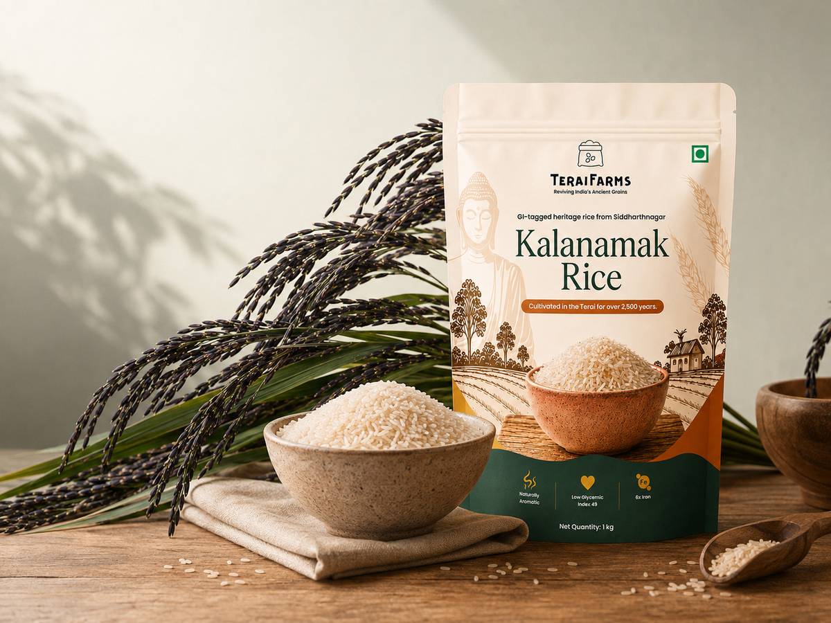 A bowl of milled short-grain Kalanamak rice beside black-husked Kalanamak paddy on green stalks and a 1 kg TeraiFarms pack, on a wooden table in soft daylight.